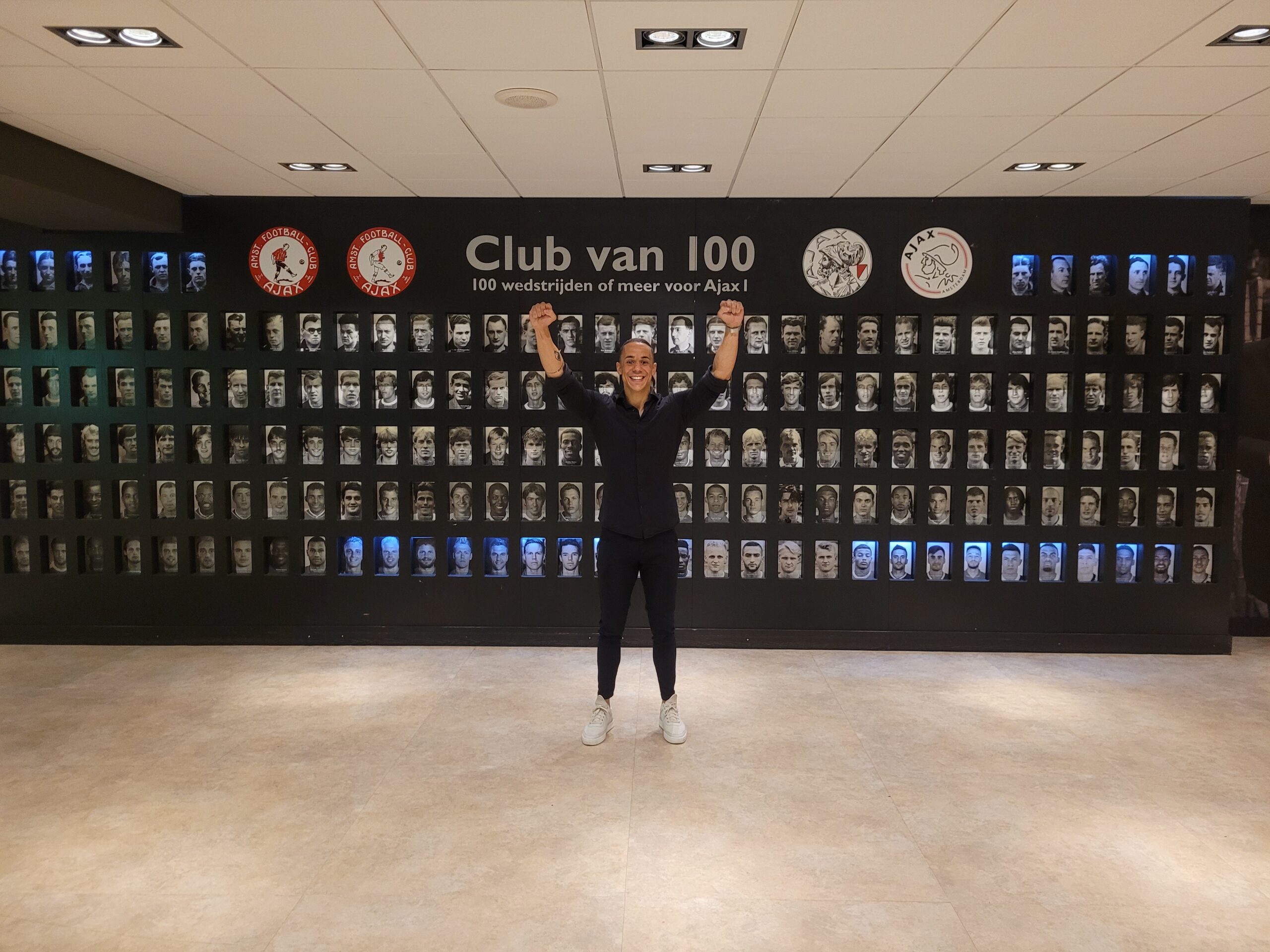 Yannick poseert juichend voor de "Club van 100" muur met portretten van Ajax-spelers in een hal.