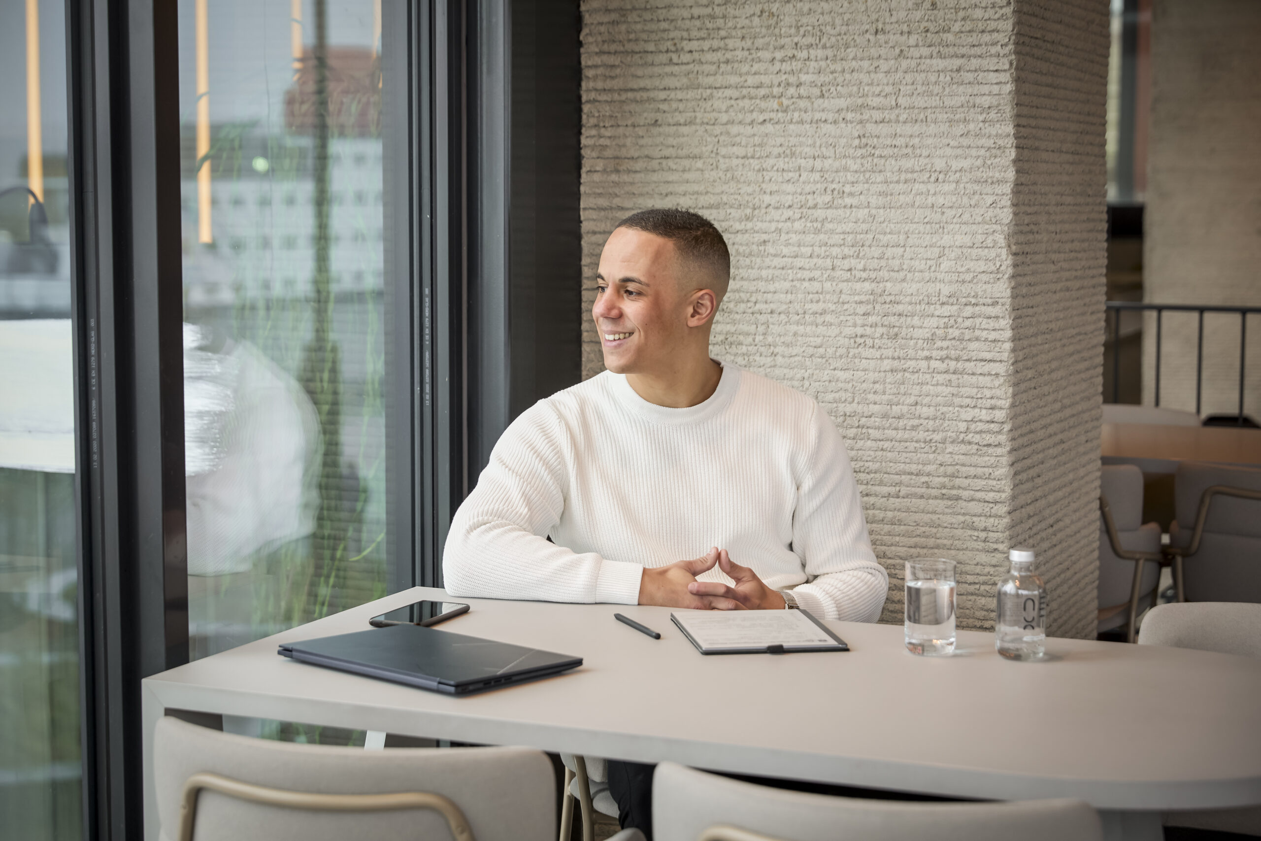 Yannick in een witte trui zit glimlachend aan een tafel in een moderne kantooromgeving met een laptop, smartphone, en een flesje water.