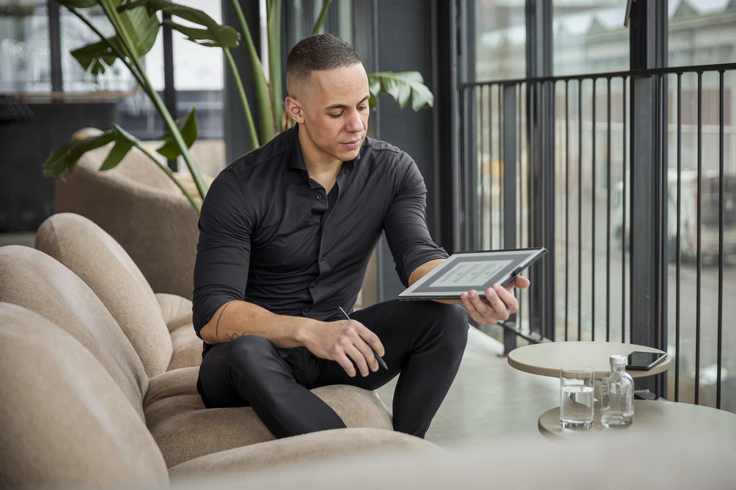 Yannick in een moderne kantooromgeving met een tablet in zijn hand, zittend op een comfortabele bank aan een tafel met glazen water.