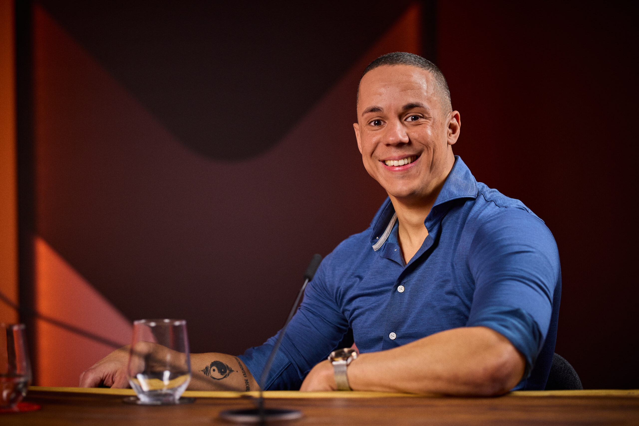 Een lachende Yannick in een blauwe overhemd zit aan een tafel met een glas water en een microfoon.