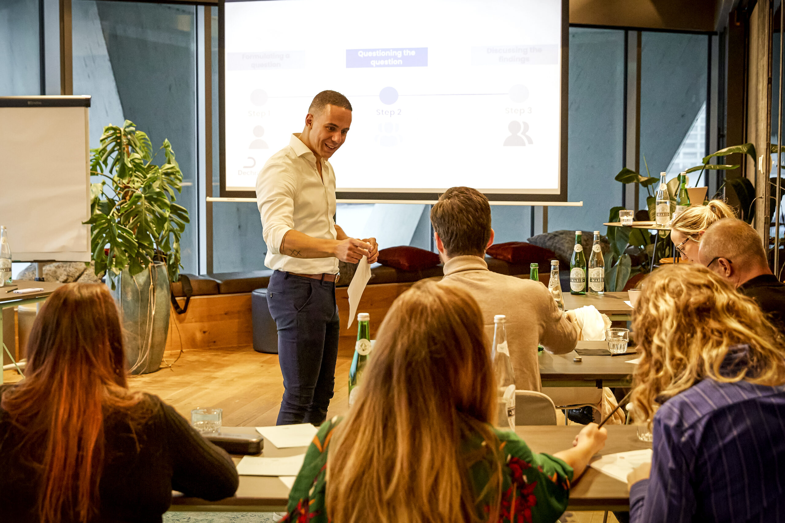 Een enthousiaste trainer, Yannick, presenteert een workshop aan een groep deelnemers in een moderne vergaderruimte.