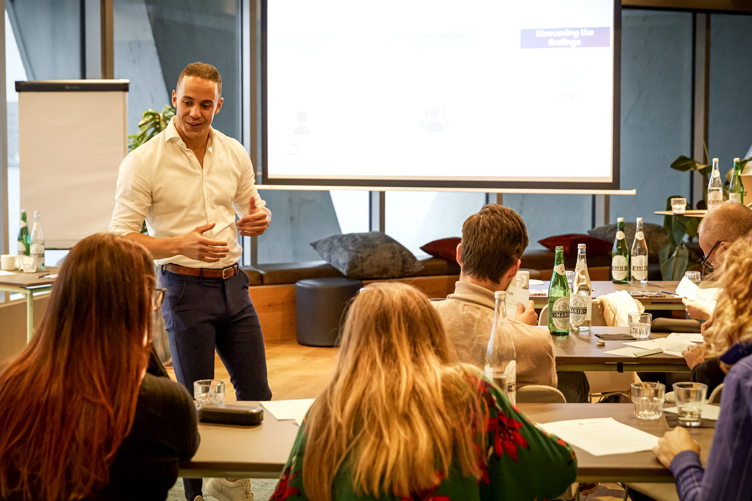 Yannick in een witte blouse geeft een presentatie aan een groep mensen in een vergaderruimte met een projectiescherm op de achtergrond.