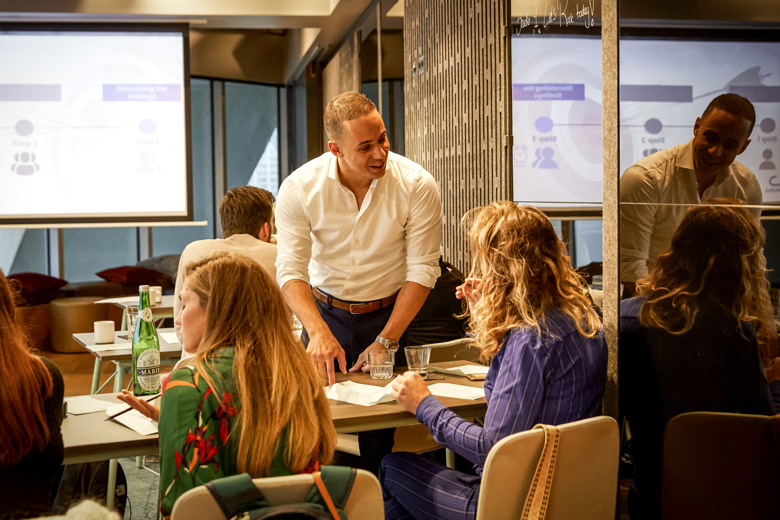 De professionele trainer, Yannick, in een witte blouse begeleidt een groep mensen tijdens een workshop, terwijl presentaties op een scherm worden weergegeven.