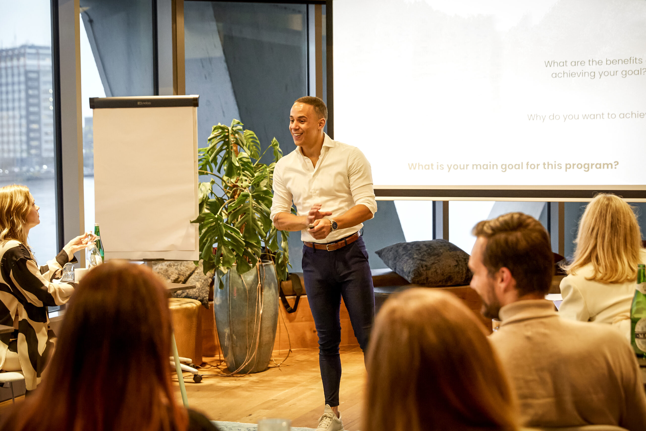 De enthousiaste spreker Yannick geeft een presentatie voor een groep mensen in een moderne vergaderruimte met een schildersezel en projectiescherm op de achtergrond.
