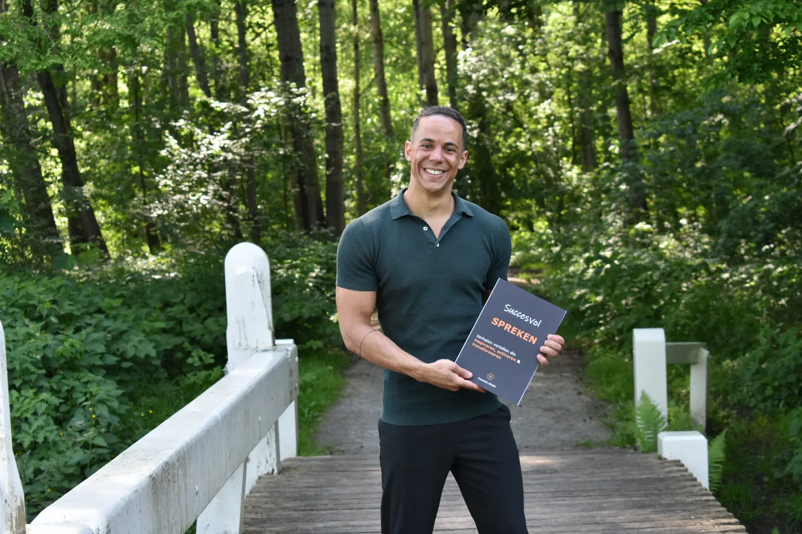 Yannick met een groen poloshirt poseert glimlachend op een houten brug in het bos met een boek getiteld "Succesvol Spreken" in zijn hand.