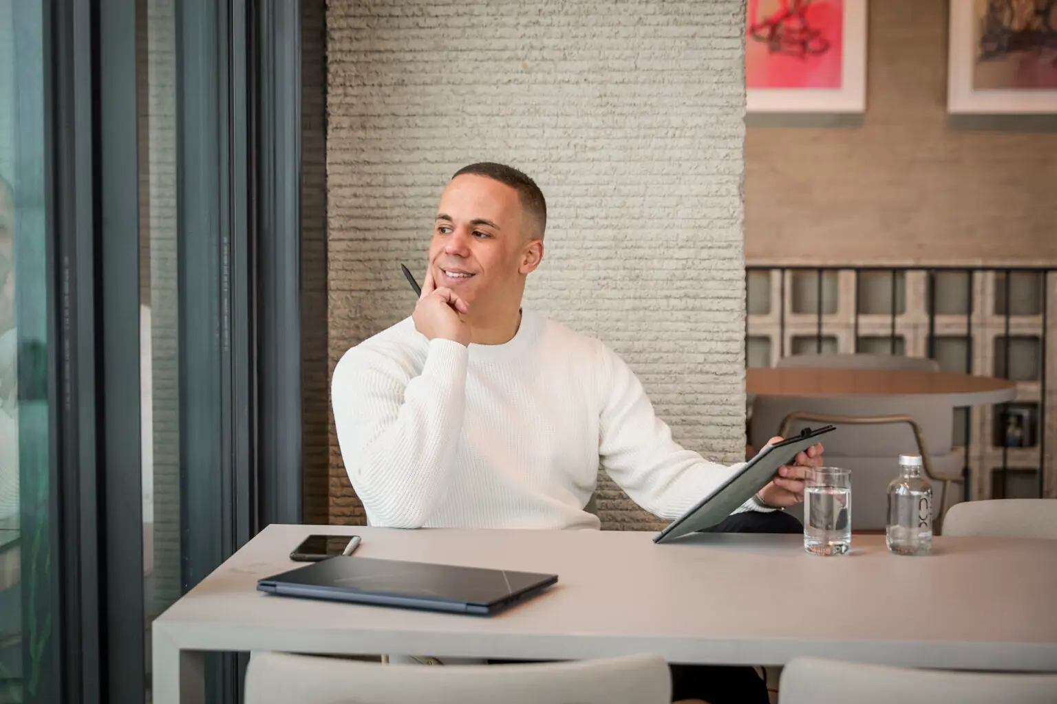 Yannick in een witte trui, zittend aan een tafel met een laptop, glas water en een notitieblok, kijkt glimlachend naar buiten.