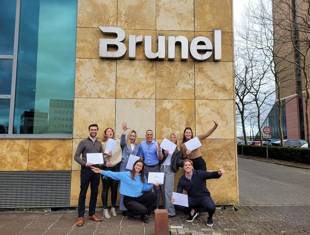 Een groep mensen poseert lachend met certificaten voor het kantoorgebouw van Brunel.