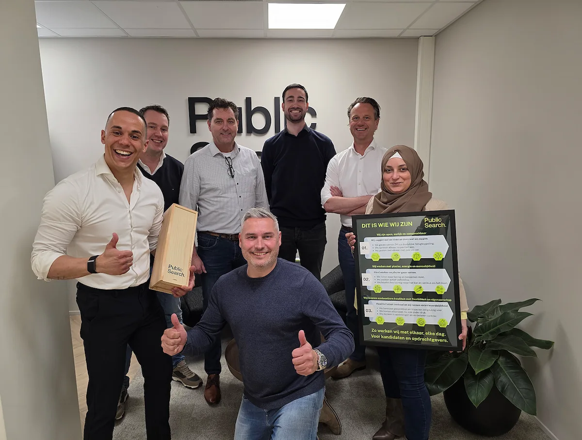 Een groep collega's van Public Search poseert vrolijk met een houten doos en een informatiebord in een kantooromgeving.
