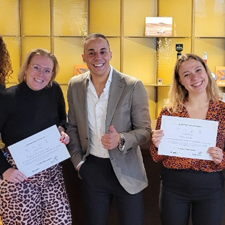 Drie mensen poseren glimlachend met certificaten voor een gele achtergrond.