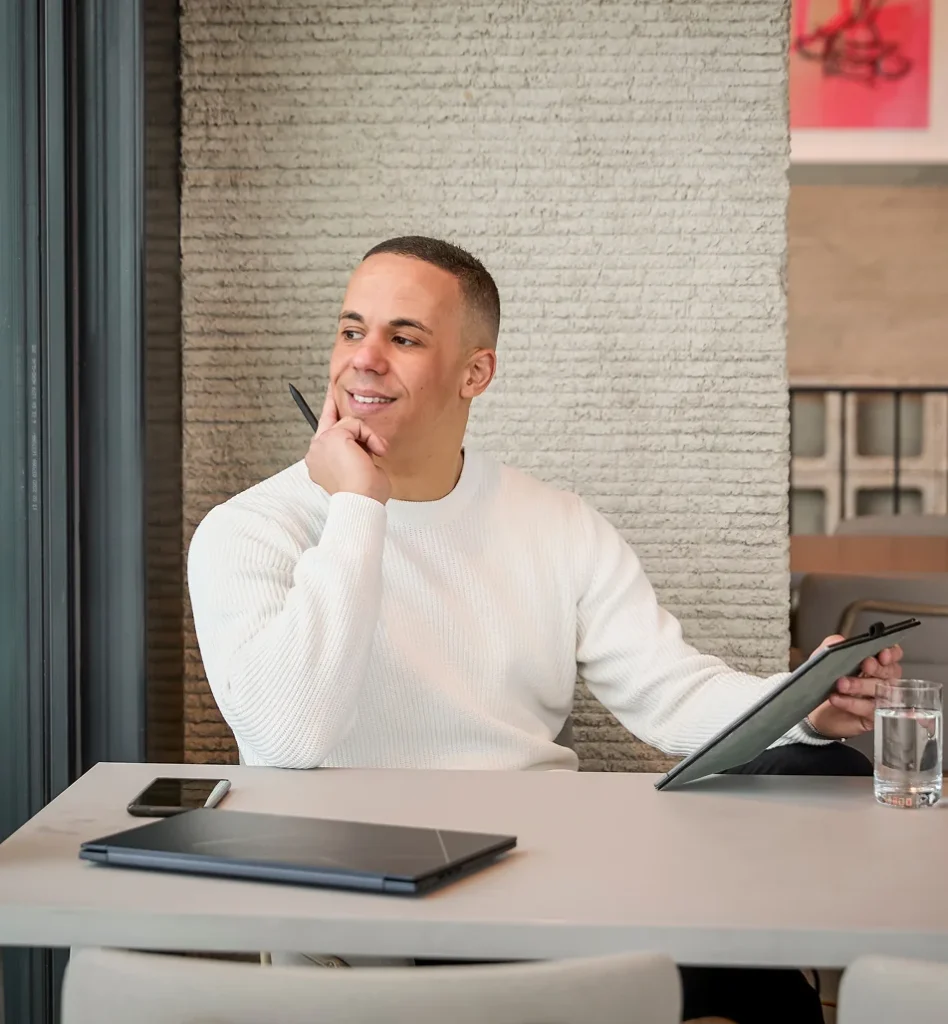 Yannick in een witte trui denkt na terwijl hij aan een tafel zit met een laptop, tablet en glas water.