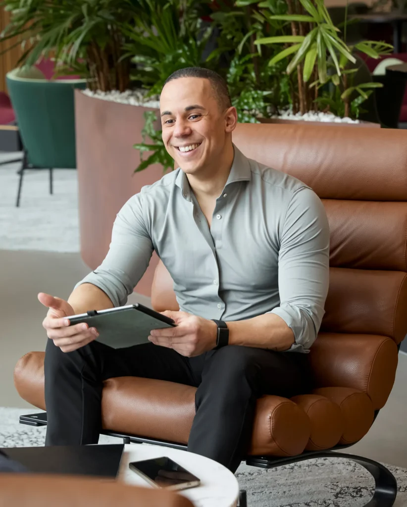 Een lachende Yannick in een moderne kantooromgeving zit ontspannen in een leren stoel met een tablet in zijn handen.