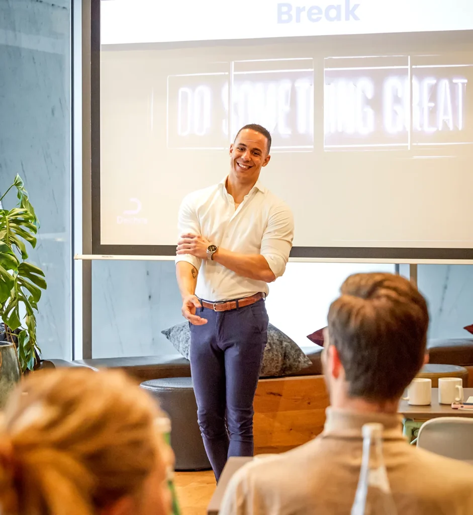 Een lachende Yannick in een witte overhemd geeft een presentatie voor een projectiescherm met de tekst "Do Something Great".