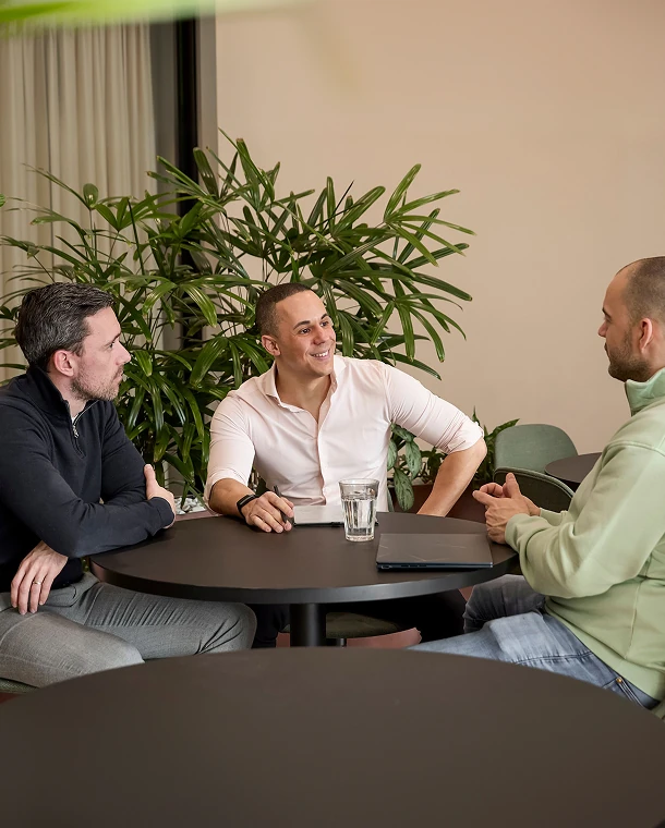 Drie mannen in gesprek aan een ronde tafel in een moderne, groene kantooromgeving.