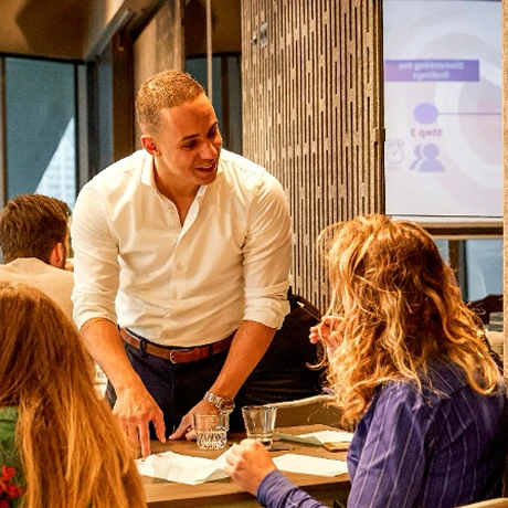 Yannick in een witte blouse geeft uitleg aan een groep mensen tijdens een bijeenkomst in een moderne vergaderruimte.